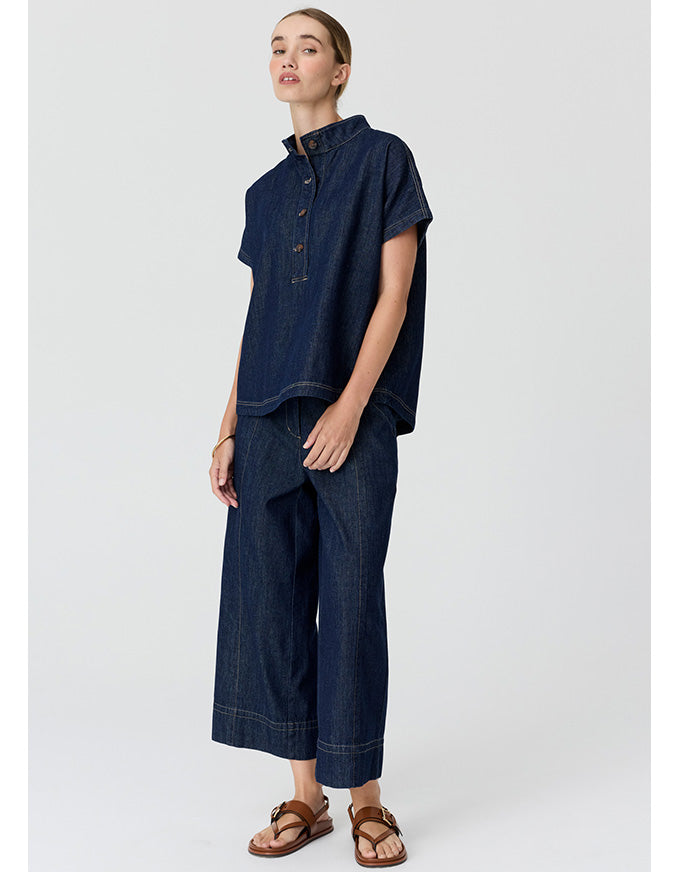 A model wearing a dark indigo denim two-piece set consisting of a short-sleeve button-up shirt and matching wide-leg cropped pants, paired with brown leather sandals, against a light gray studio background.