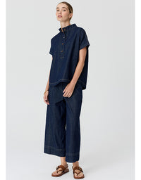 A model wearing a dark indigo denim two-piece set consisting of a short-sleeve button-up shirt and matching wide-leg cropped pants, paired with brown leather sandals, against a light gray studio background.