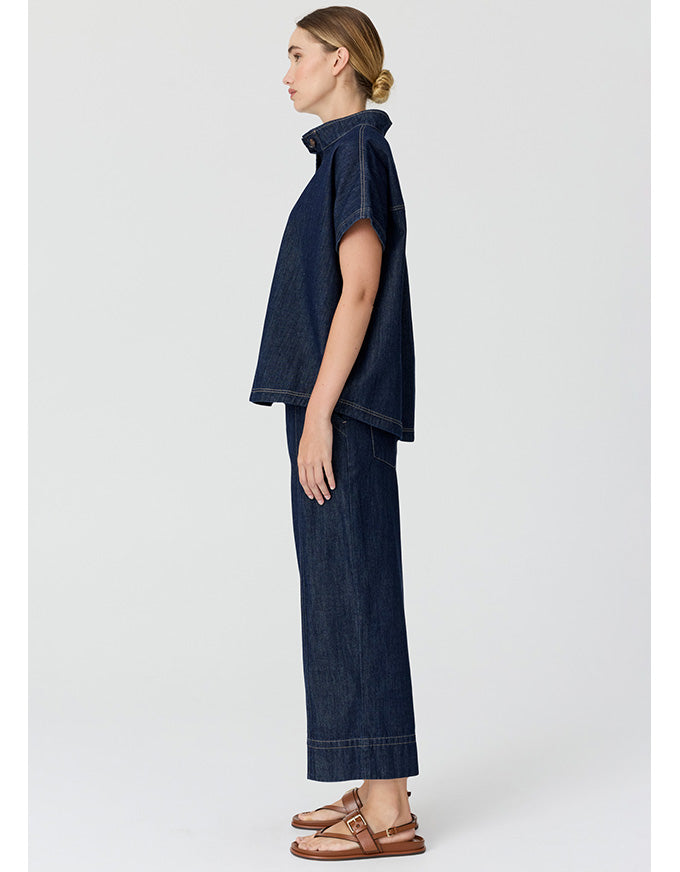 A model shown in profile wearing a dark indigo denim two-piece set consisting of a short-sleeve cropped top and matching wide-leg trousers, paired with brown leather sandals, against a light gray studio background.