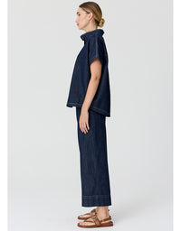 A model shown in profile wearing a dark indigo denim two-piece set consisting of a short-sleeve cropped top and matching wide-leg trousers, paired with brown leather sandals, against a light gray studio background.