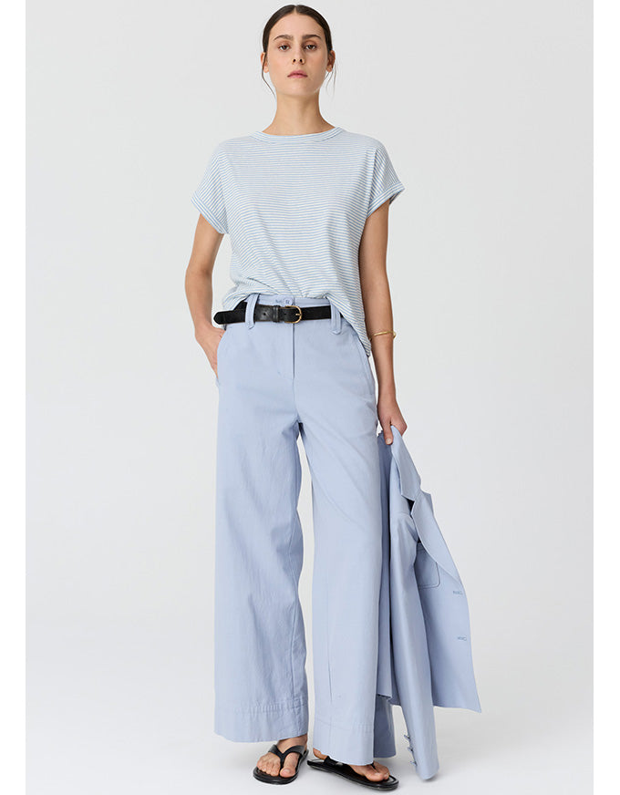 A woman stands against a light gray studio background wearing a light blue short-sleeve striped T-shirt, light blue wide-leg pants, a black belt, and black sandals, while holding a light blue blazer.