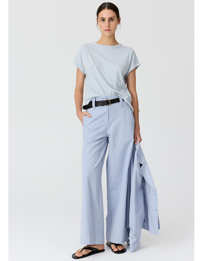 A woman modeling a light blue and white striped short-sleeve t-shirt, light blue wide-leg trousers, a black belt, and black sandals, holding a light blue blazer against a plain light gray studio background.