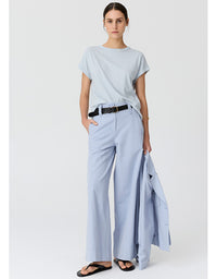 A woman modeling a light blue and white striped short-sleeve t-shirt, light blue wide-leg trousers, a black belt, and black sandals, holding a light blue blazer against a plain light gray studio background.