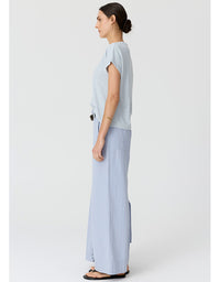 A woman shown in profile wearing a light blue and white striped short-sleeve top with matching wide-leg light blue pants, standing against a plain light gray studio background.