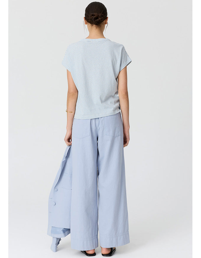 A model shown from the back wearing a light blue and white striped short-sleeve t-shirt, light blue wide-leg trousers, and holding a matching light blue jacket against a plain light gray studio background.
