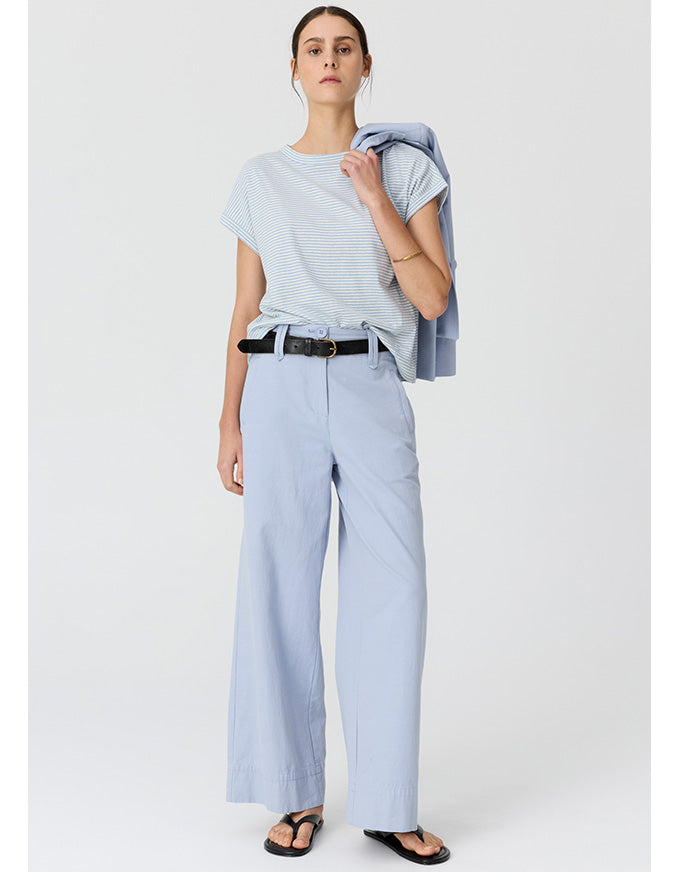A woman modeling a light blue short-sleeve striped t-shirt, matching wide-leg trousers, and black sandals, with a light blue blazer draped over her shoulder, against a plain light gray studio background.