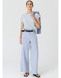 A woman modeling a light blue short-sleeve striped t-shirt, matching wide-leg trousers, and black sandals, with a light blue blazer draped over her shoulder, against a plain light gray studio background.