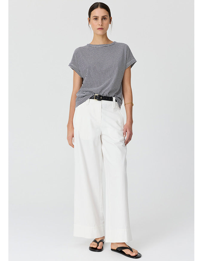 A woman modeling a short-sleeve gray and white striped t-shirt, paired with white wide-leg trousers and black sandals, against a light gray studio background.