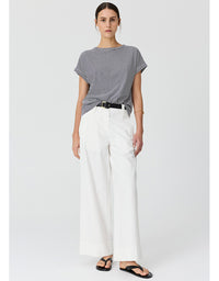 A woman modeling a short-sleeve gray and white striped t-shirt, paired with white wide-leg trousers and black sandals, against a light gray studio background.