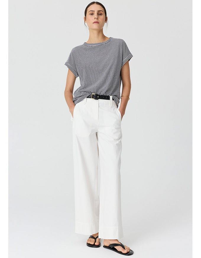 A woman modeling a short-sleeve gray and white striped t-shirt paired with white wide-leg trousers and black sandals, standing against a light gray studio background.
