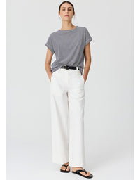 A woman modeling a short-sleeve gray and white striped t-shirt paired with white wide-leg trousers and black sandals, standing against a light gray studio background.