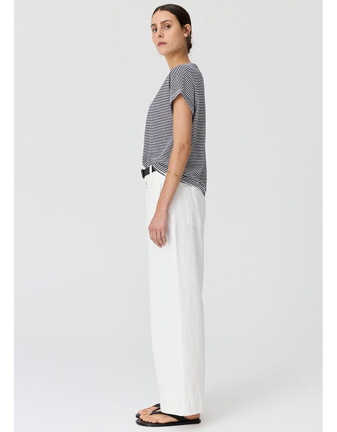 A woman stands in profile wearing a short-sleeve black and white striped top, white wide-leg trousers, and black flip-flops against a light gray studio background.