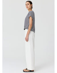 A woman stands in profile wearing a short-sleeve black and white striped top, white wide-leg trousers, and black flip-flops against a light gray studio background.
