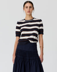 A model wearing a short-sleeve knit top with horizontal navy and off-white stripes, paired with a dark navy skirt, against a light gray studio background.
