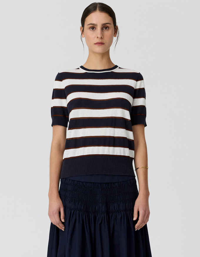 A woman modeling a short-sleeve knit top with horizontal navy and off-white stripes, paired with a dark navy skirt, against a light gray studio background.
