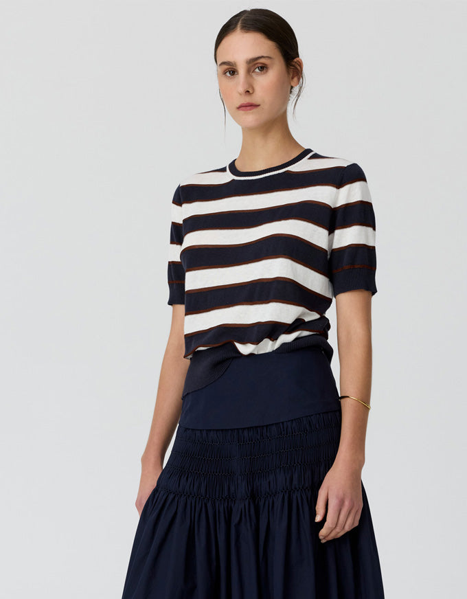 A woman modeling a short-sleeve knit top with horizontal navy and off-white stripes, paired with a dark navy skirt, against a light gray studio background.