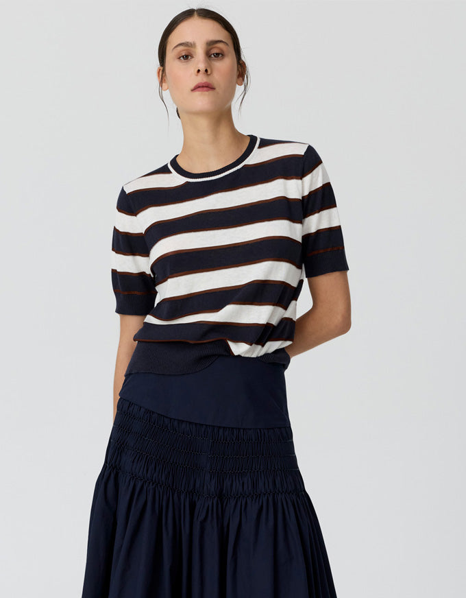 A woman modeling a short-sleeve knit top with horizontal navy and off-white stripes, paired with a dark navy skirt, against a light gray studio background.