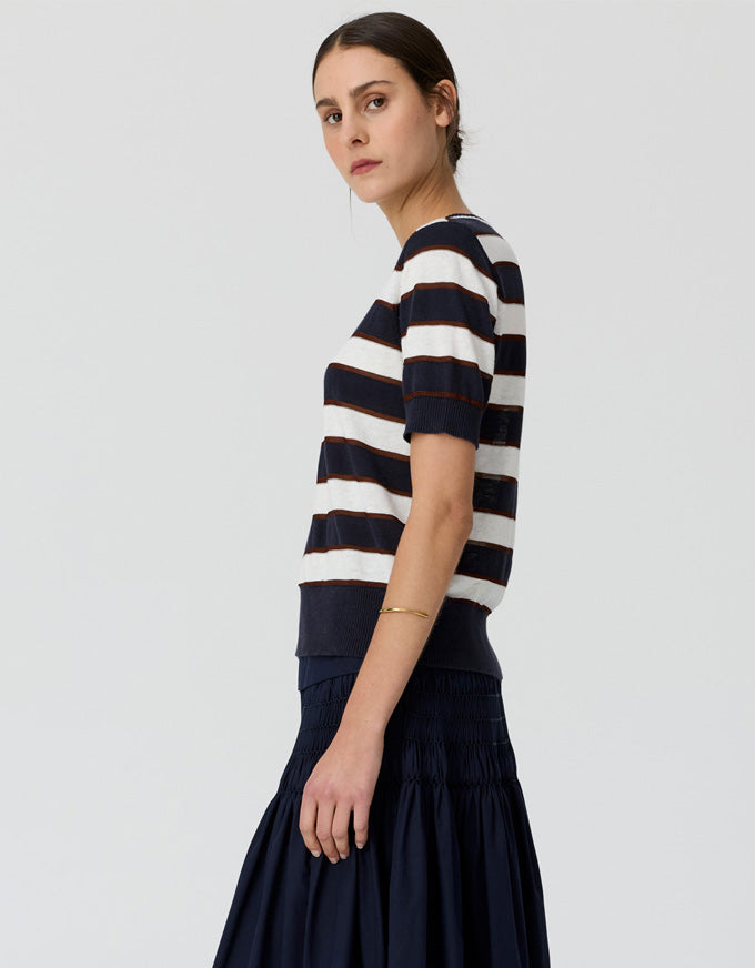 A woman shown in side profile wearing a short-sleeve knit top with horizontal navy and off-white stripes, paired with a dark navy skirt, against a light gray studio background.