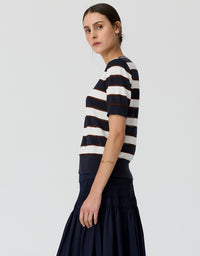 A woman shown in side profile wearing a short-sleeve knit top with horizontal navy and off-white stripes, paired with a dark navy skirt, against a light gray studio background.