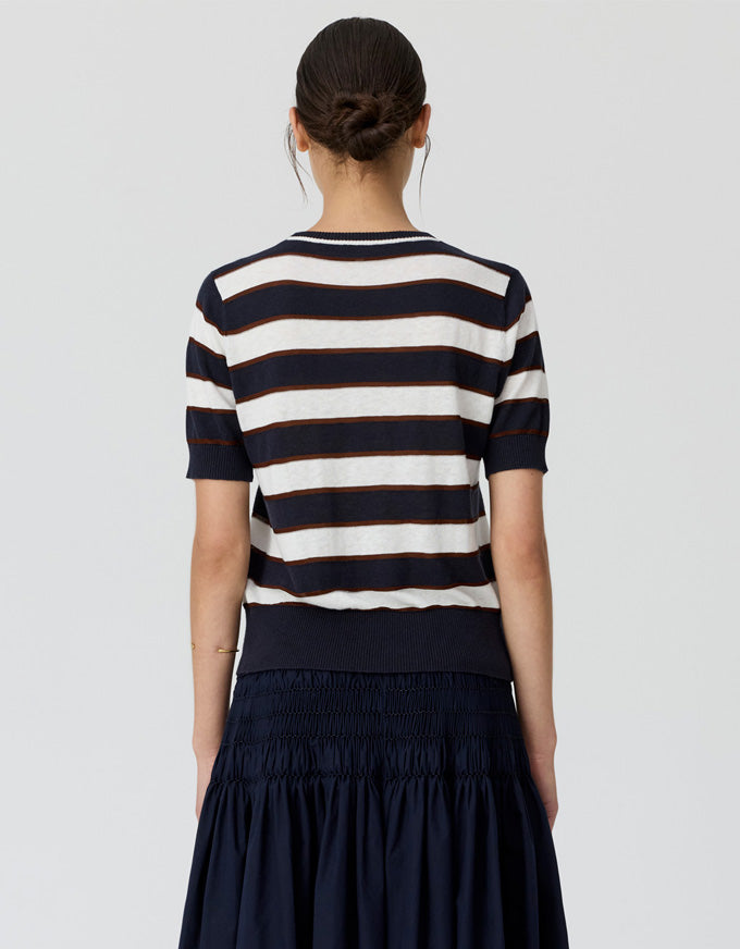 A woman viewed from the back wearing a short-sleeve knit top with horizontal navy and off-white stripes, paired with a dark navy skirt, against a light gray studio background.