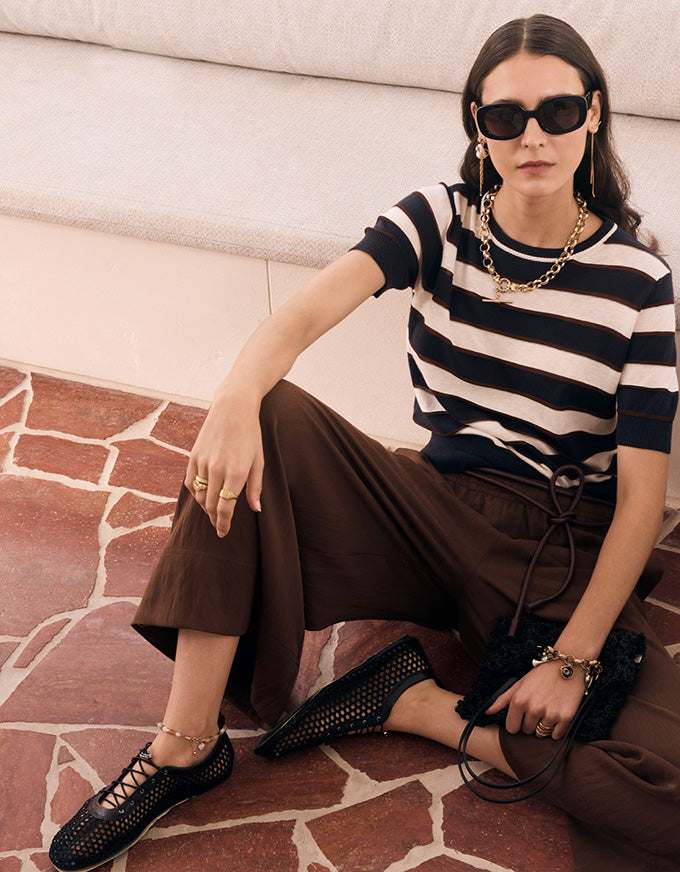 A woman sits on a terracotta stone floor in front of a cream-colored sofa, wearing a black and white horizontally striped short-sleeve knit top, brown wide-leg trousers, black perforated lace-up flats, gold jewelry, and black sunglasses.