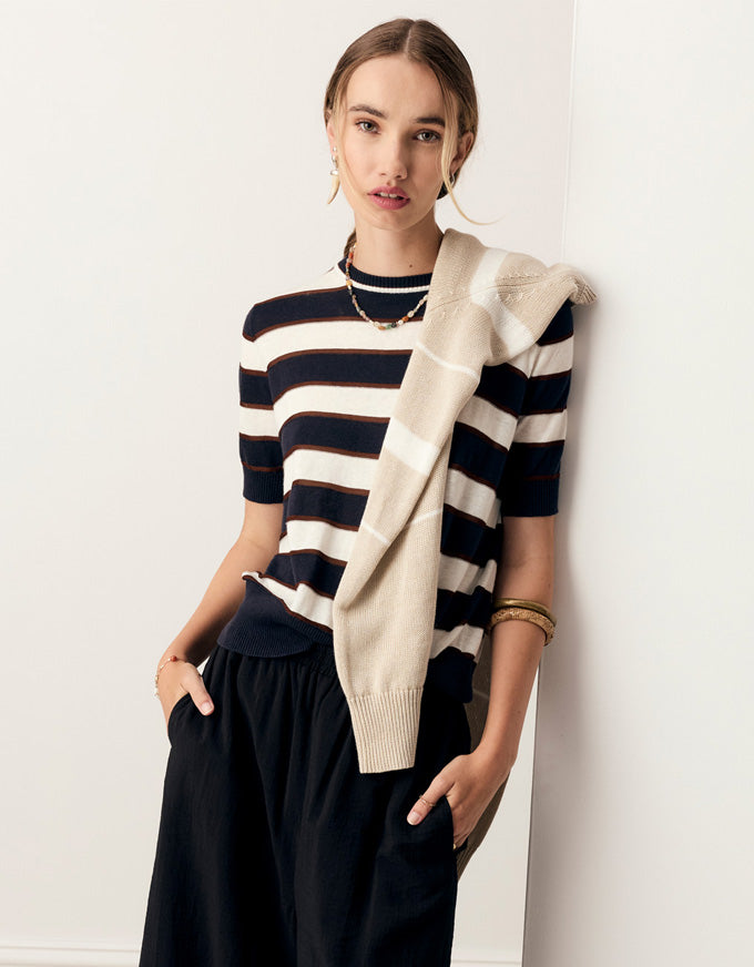 A woman models a short-sleeve knit top with navy and cream horizontal stripes, styled with a cream knit cardigan draped over her shoulder and dark wide-leg pants, set against a plain white wall.