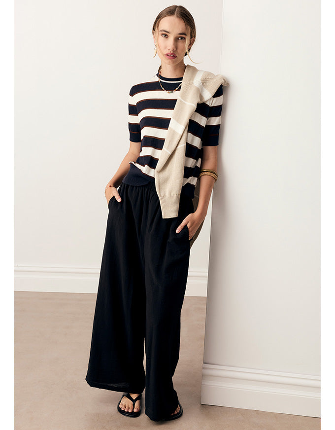 A woman stands against a white wall wearing a navy and cream striped short-sleeve knit top, black wide-leg pants, and black sandals, with a cream knit wrap draped over one shoulder.