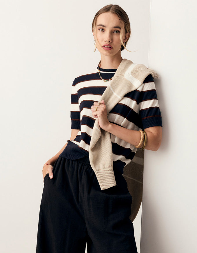 A woman models a navy and cream striped short-sleeve knit top, paired with dark wide-leg trousers and holding a cream knit cardigan against a clean white wall.