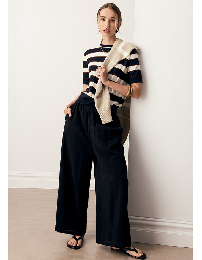 A woman models a black and white striped short-sleeve top with wide-leg black pants and a beige knit cardigan draped over her shoulder, standing against a white wall.