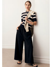 A woman models a black and white striped short-sleeve top with wide-leg black pants and a beige knit cardigan draped over her shoulder, standing against a white wall.