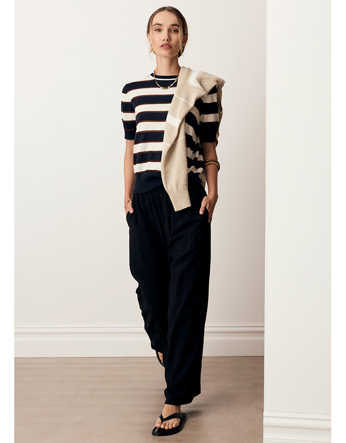 A woman models a short-sleeve knit top with navy and cream horizontal stripes, paired with loose black pants and a draped cream cardigan, in a minimalist indoor setting with a white wall and light wood floor.