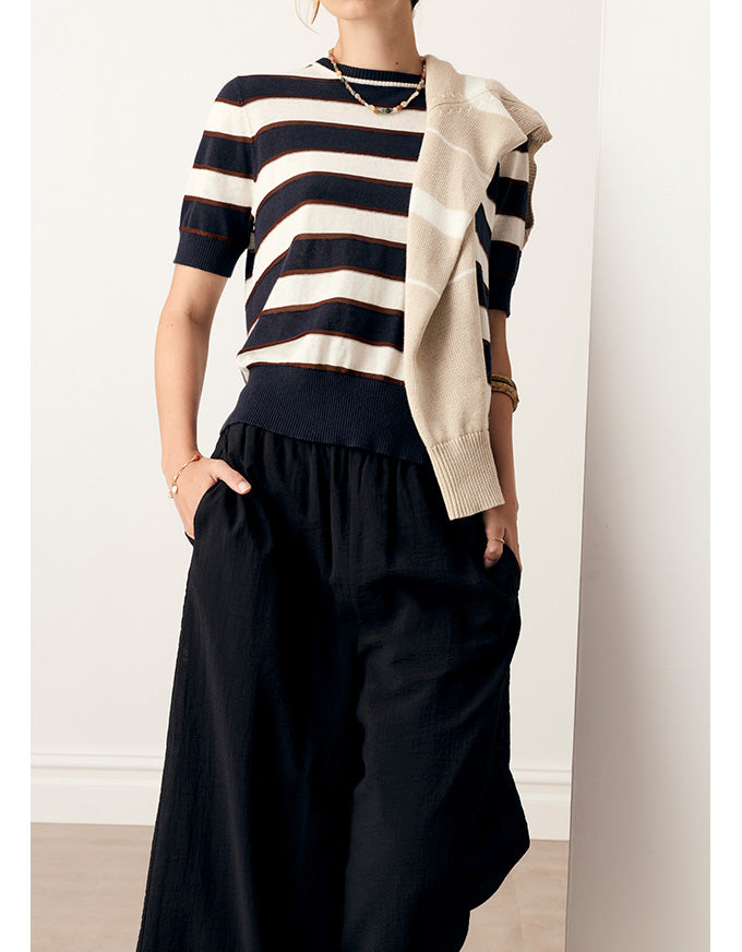 A model wearing a short-sleeve knit top with horizontal navy and cream stripes, black wide-leg pants, and a draped cream cardigan, set against a minimalist off-white wall.
