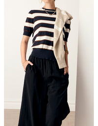 A model wearing a short-sleeve knit top with horizontal navy and cream stripes, black wide-leg pants, and a draped cream cardigan, set against a minimalist off-white wall.