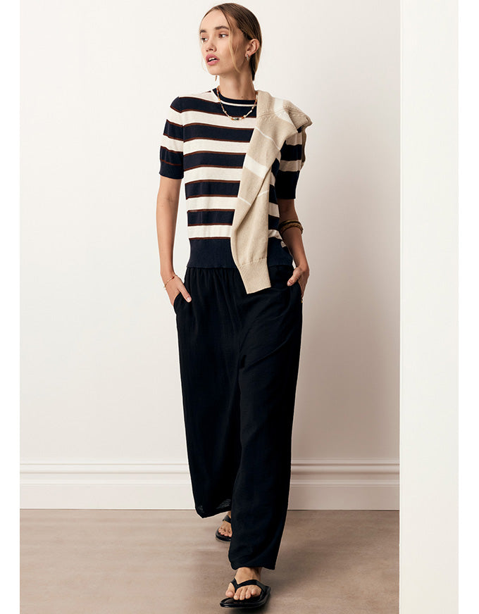 A woman models a short-sleeve knit top with navy and cream horizontal stripes, styled with a draped cream cardigan, loose dark navy pants, and black sandals against a neutral indoor wall.