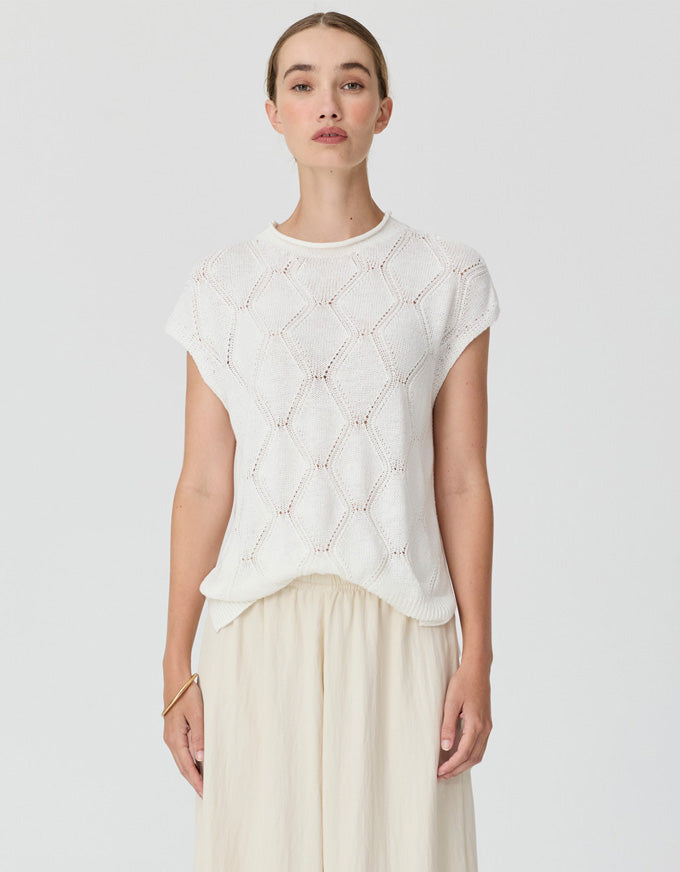 A woman modeling a short-sleeve white knit top with a textured geometric diamond pattern, paired with light beige wide-leg trousers, against a solid light gray background.