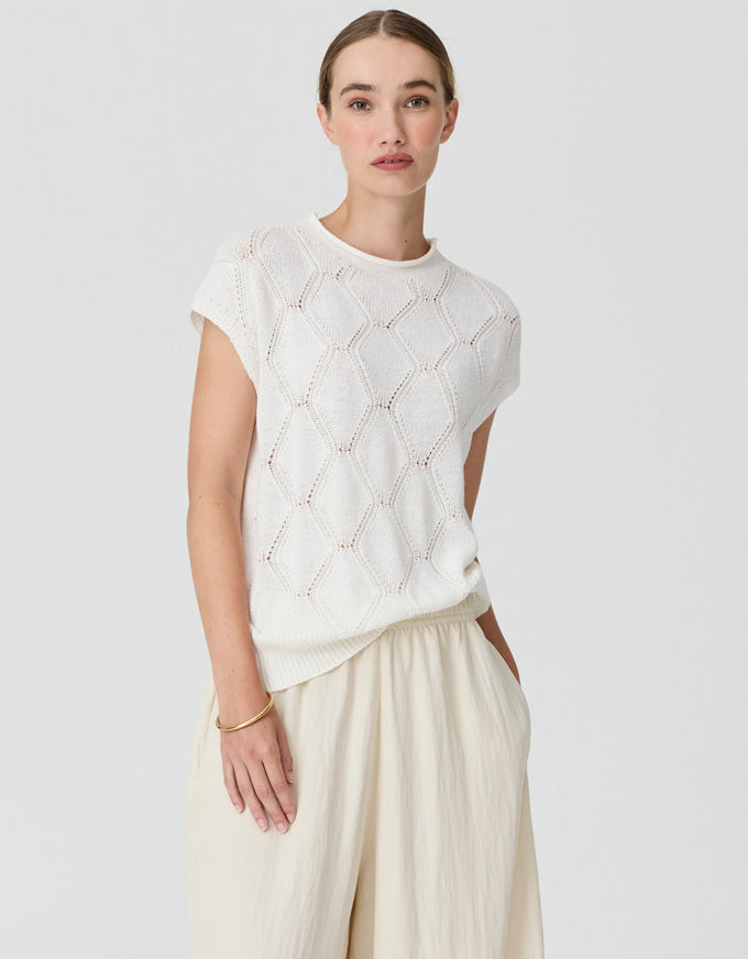 A woman modeling a short-sleeve cream knit top with a textured openwork diamond pattern, paired with cream wide-leg trousers, against a light gray studio background.