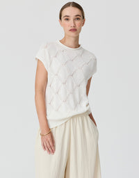 A woman modeling a short-sleeve cream knit top with a textured openwork diamond pattern, paired with cream wide-leg trousers, against a light gray studio background.