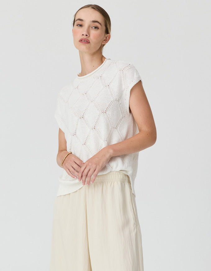 A woman modeling a white sleeveless knit top with a textured geometric pattern, paired with light beige wide-leg trousers, against a solid light gray background.
