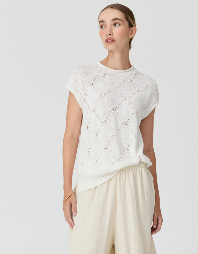 A woman wearing a white short-sleeve knit top with a textured openwork diamond pattern and cream wide-leg pants, photographed against a light gray studio background.