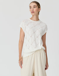 A woman wearing a white short-sleeve knit top with a textured openwork diamond pattern and cream wide-leg pants, photographed against a light gray studio background.