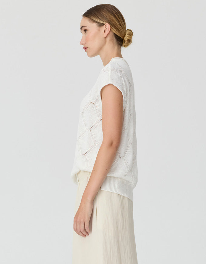 A woman shown in side profile wearing a white sleeveless knit vest over a cream top and light beige skirt, photographed against a plain light gray studio background.