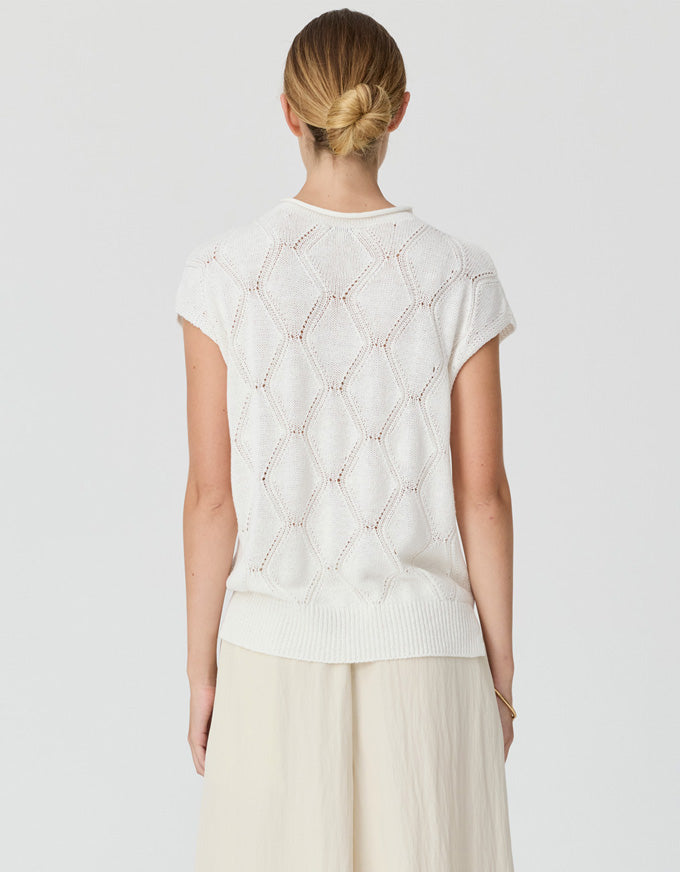 A woman viewed from behind wearing a cream-colored short-sleeve knit top with a textured geometric pattern and light beige wide-leg trousers, set against a plain light gray studio background.