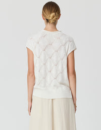 A woman viewed from behind wearing a cream-colored short-sleeve knit top with a textured geometric pattern and light beige wide-leg trousers, set against a plain light gray studio background.
