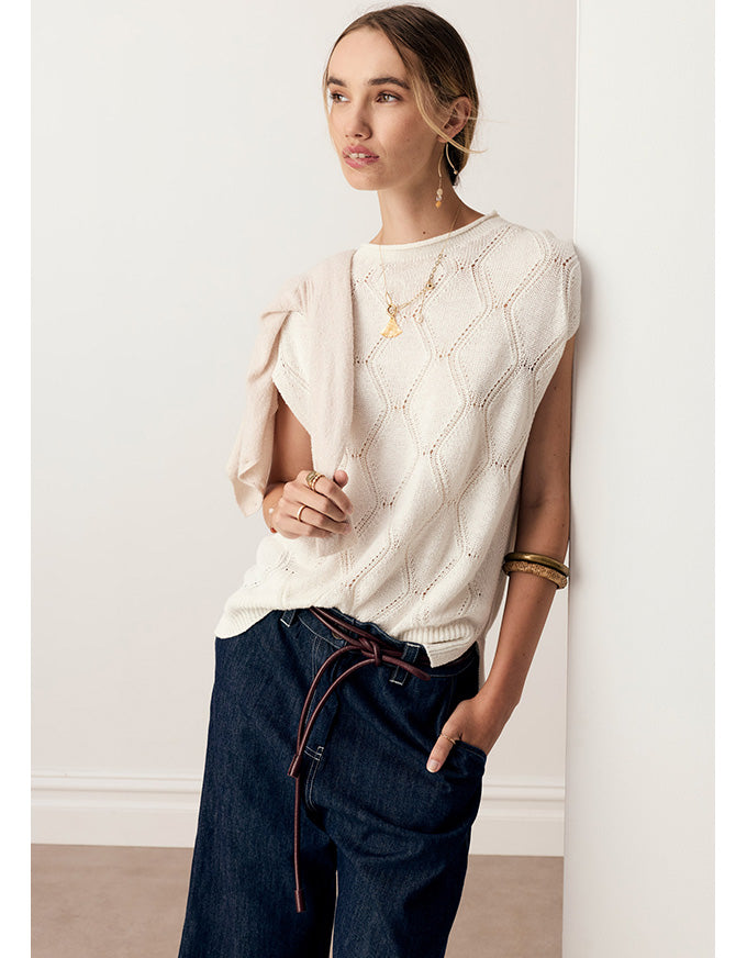 A woman models a cream-colored sleeveless knit top with textured diamond patterns and draped puff sleeves, paired with dark blue jeans, standing against a clean white wall.
