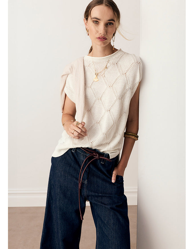 A woman modeling a cream-colored sleeveless knit top with a textured geometric pattern, paired with dark wide-leg jeans featuring a brown drawstring, standing against a plain off-white wall.