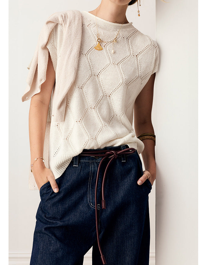 A person wearing a cream-colored sleeveless knit sweater with a geometric openwork pattern, paired with dark blue jeans and gold jewelry, standing against a white wall.