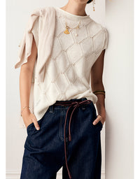 A person wearing a cream-colored sleeveless knit sweater with a geometric openwork pattern, paired with dark blue jeans and gold jewelry, standing against a white wall.