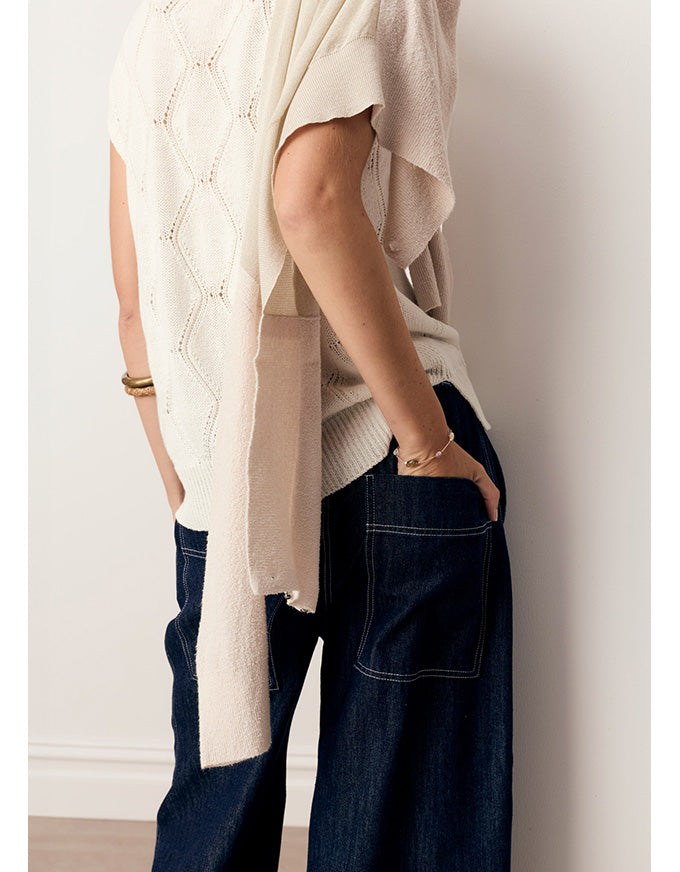 A person wearing a cream-colored open-knit draped cardigan over a ribbed knit top and dark blue wide-leg jeans, standing against a plain off-white wall.