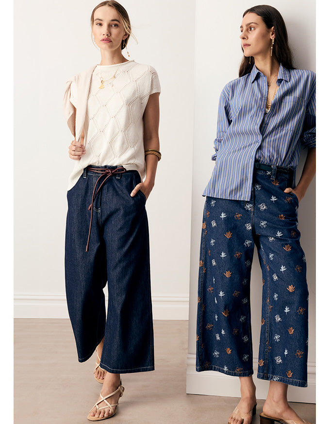 Two women modeling wide-leg cropped pants in a bright indoor setting. The left model wears dark blue wide-leg denim pants with a drawstring waist and a cream knit top.