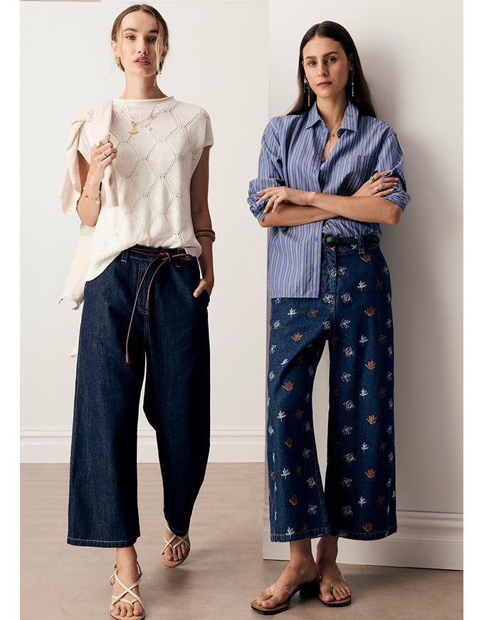 Two women modeling wide-leg pants in a minimalist indoor setting. The woman on the left wears dark blue denim wide-leg pants with a brown drawstring waist and a cream textured top.